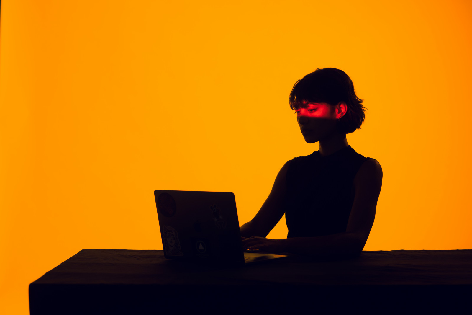A lone figure silhouetted at a laptop, lit by a single band of red light.